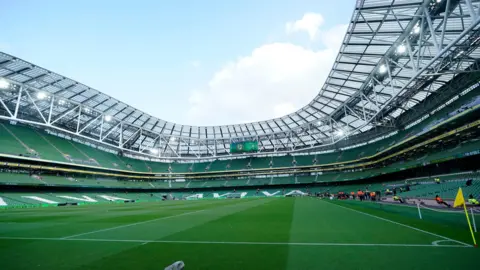 PA An empty Aviva Stadium in Dublin, shown when no concerts or sporting events are taking place. Three quarters the stadium and three tiers of green seats can be seen surrounding a lush football pitch. 