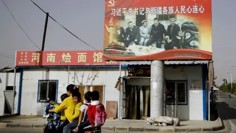 AP Photo/Andy Wong A Uyghur woman uses an electric-powered scooter to fetch school children as they ride past a picture showing China's President Xi Jinping joining hands with a group of Uighur elders at the Unity New Village in Hotan, in western China's Xinjiang region.