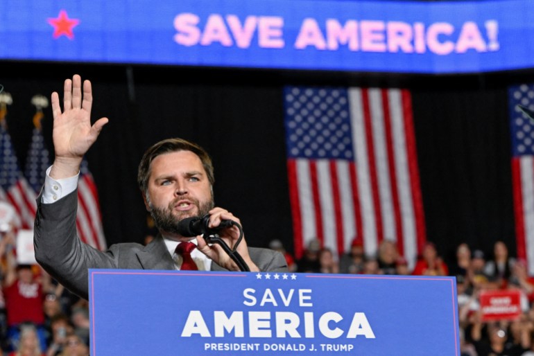 JD Vance raises a hand as he speaks behind a podium that reads, "Save America." 