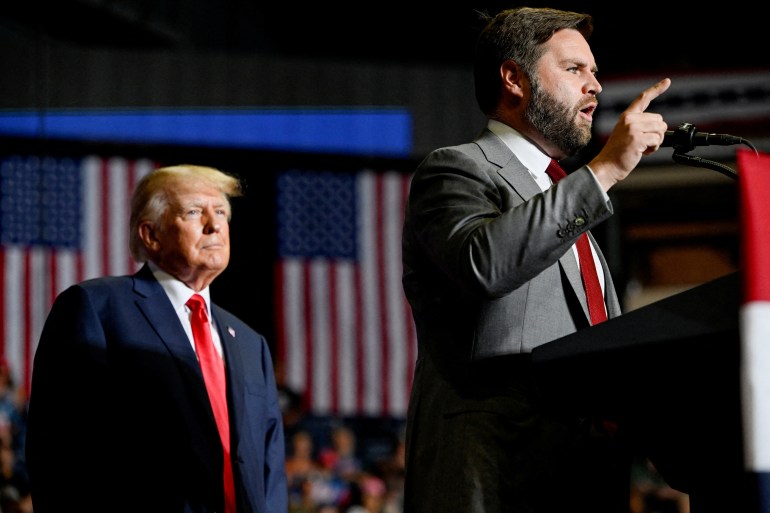 Donald Trump stands behind JD Vance, who speaks at a podium with finger pointed.