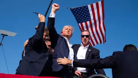 AP Donald Trump with blood on his face, surrounded by staff in front of an American flag