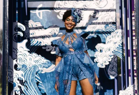 RAJESH JANTILAL/AFP A woman wears denim-blue clothing in front of a blue stage set. She is smiling.