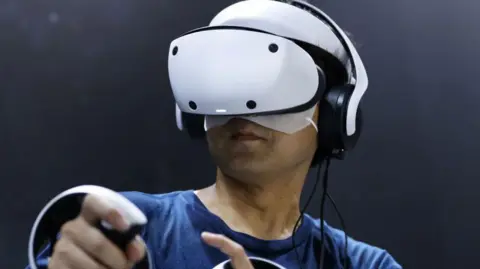 Getty Images A man uses the PlayStation VR 2. It is larger than the other two headsets, and it is a sleek white curved shape.