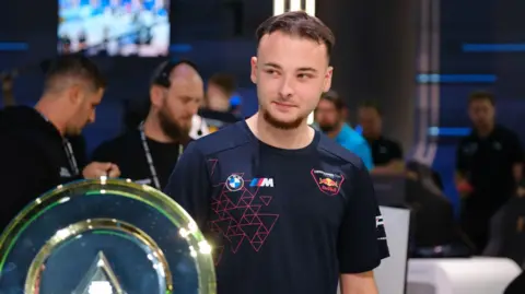 Esports World Cup Foundation Luke Bennett, pictured with a trophy at the Esports World Cup in Saudi Arabia.  Luke is a 19-year-old man with short brown hair and a short goatee beard. He wears a black T-shirt. 