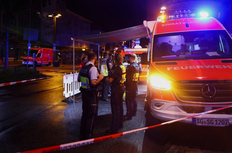 Police vehicles with lights blazing illuminate an area cordoned off by crime-scene tape, where officers look around.