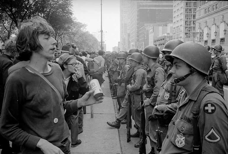 An archival, black-and-white photo shows armed, helmeted police and demonstrators lined up, facing each other on a broad Chicago avenue.