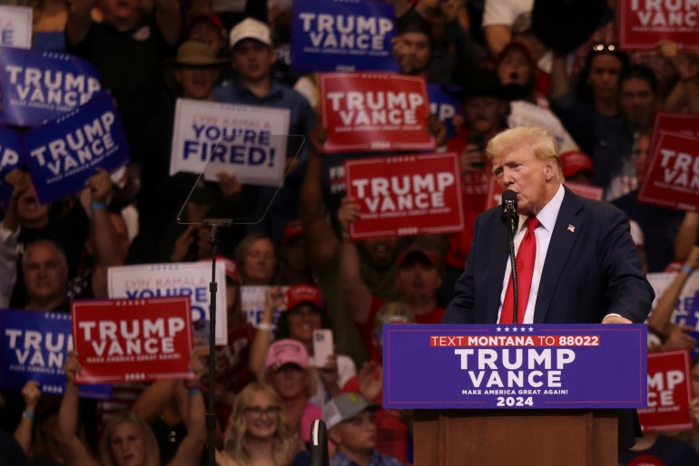 Donald Trump speaks into a microphone at a podium labeled, "Trump Vance." Around him, supporters wave campaign signs.