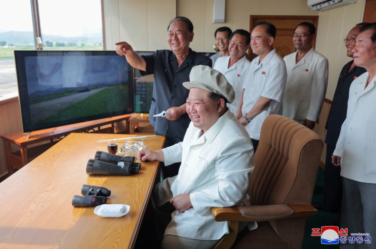 Kim Jong Un sitting at a desk. There is a screen in front of him, showing a picture of a tank. There are two pairs of binoculars on the desk. Kim is wearing a white lab coat and a puffy light beige cap on his head. He is laughing. A group of older men are with him. They are also smiling. One is pointing.