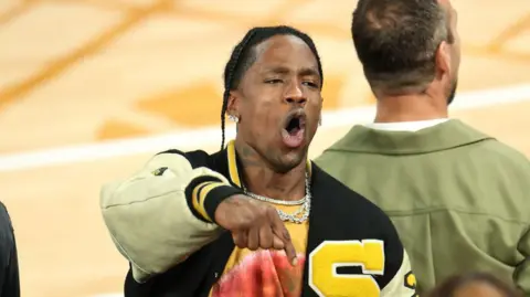 Getty Images Travis Scott wears a black and yellow varsity jacket with a large yellow "S" on the left breast. He's gesturing and shouting, pointing downwards, in an apparent celebration.