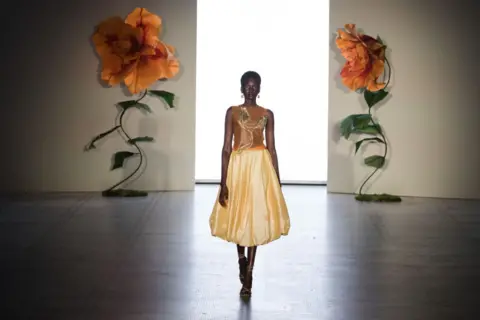 WIKTOR SZYMANOWICZ / GETTY IMAGES A model walks down a runway wearing a dress with a flower motif. The walls behind her also have two large flower motifs.