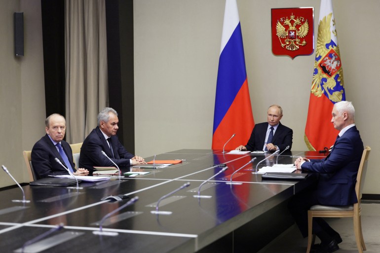 Vladimir Putin in a meeting with government officials. They are seated at a table. Putin is at the head. There are flags behind him and a coat of arms on the wall.