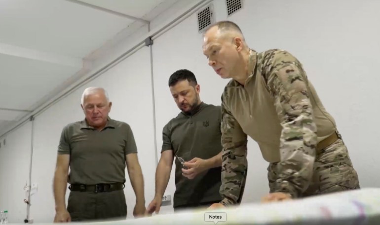 Volodymyr Zelenskyy discussing the war with his top commanders. They are standing over a desk and looking at a map