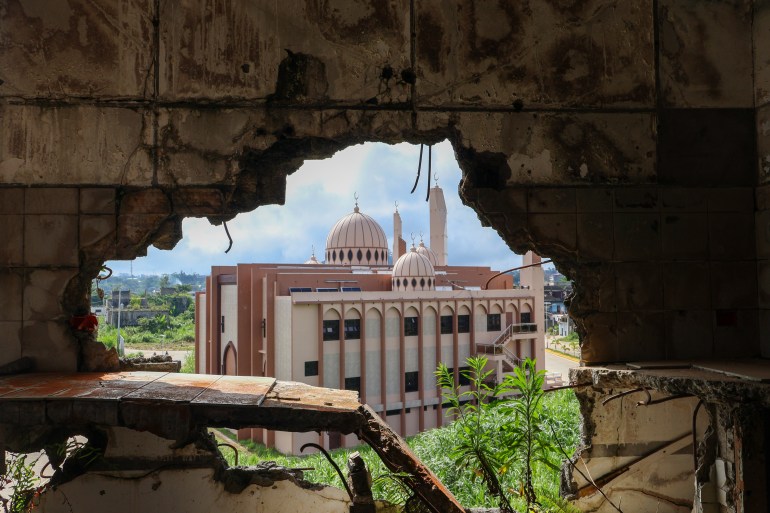 A rebuilt mosque seen through a ruined building. There are bullet holes in the all of the building