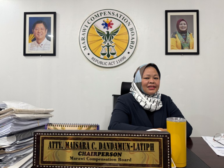 Maisara Dandamun-Latiph, She is seated at a desk with an official seals and portrait of the president on the wall behind her. She is wearing a black dress and a white headscarf.