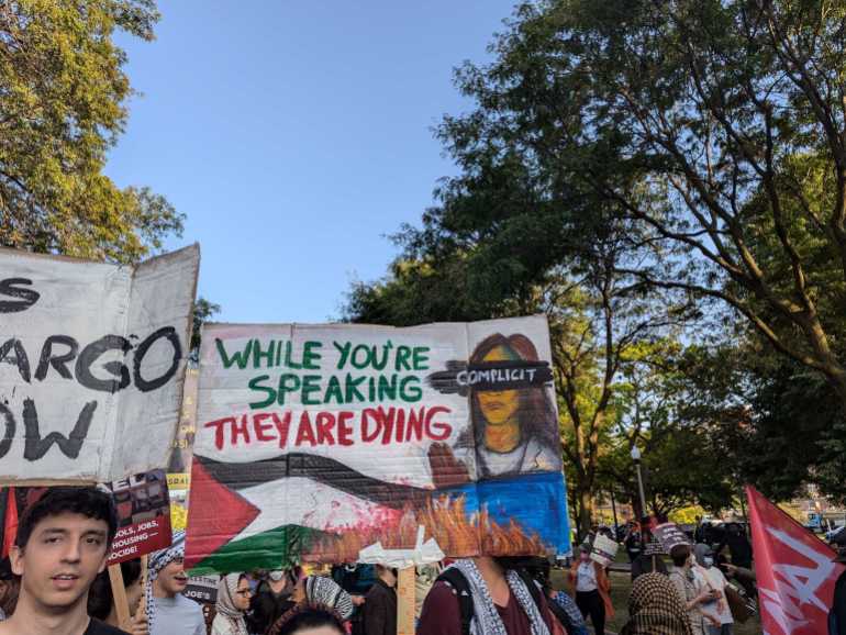 Protesters hold up hand-painted signs that read, "While you're speaking, they are dying."