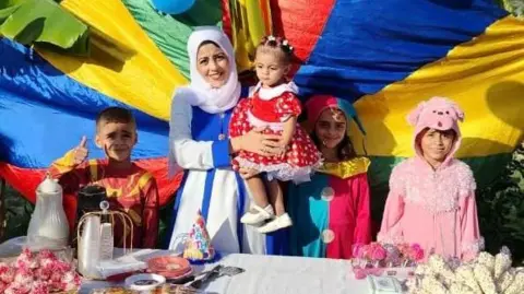 Handout Woman standing with four children - all wearing colourful clothing - standing in front of rainbow coloured cloth and behind a table that is filled with party food