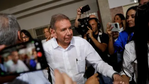 Reuters Edmundo González walks passed a number of people holding cameras. He has short, brown hair and is wearing a white shirt with a pen sticking out of his front pocket.