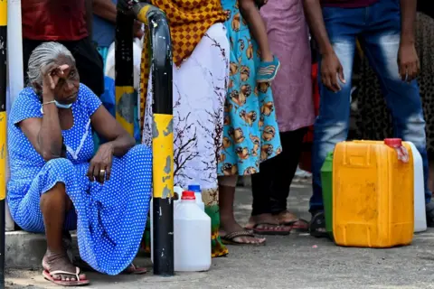 Getty Images A woman in a blue spotted dress sits in a queue of people next to gasoline containers