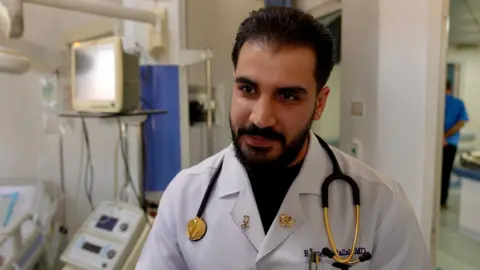 Goktay Koraltan / BBC Dr Basil Abdallah has dark hair and a beard - he is standing in a lab coat in a hospital with a gold coloured stethoscope around his neck