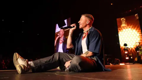 Getty Images Macklemore sits down on stage, smiling, holding a microphone up to his face. There is also an image of him on a screen behind him