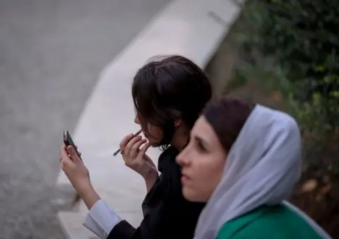 Getty Images Two women sitting at a park bench. One is wearing a headscarf, the other isn't.