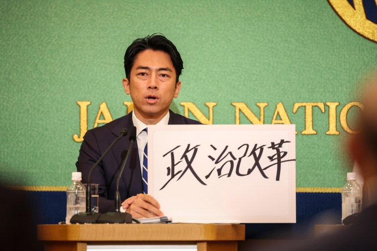 Shinjiro Koizumi. He is seated at a desk. He is holding a handwritten placard with 'Poltiical Reform' written on it in Jaoanese