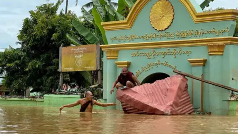 Getty Images A Buddhist monk wades through flood waters as another sits on a broken roof in front of a monastery in Sin Thay village in Pyinmana, in Myanmar's Naypyidaw region, on September 13, 2024, following heavy rains in the aftermath of Typhoon Yagi.