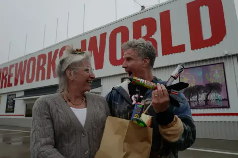 Netflix Harper Steele,  wearing a white top and taupe cardigan, holding a bag filled with fireworks alongside Will Ferrell, who is holding some firework rockets, wearing a blue jacket.