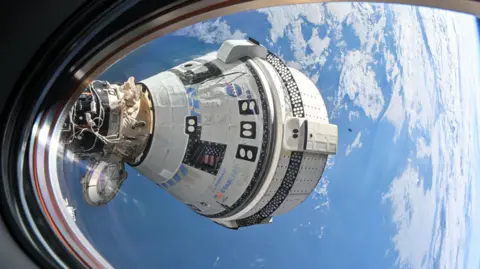 NASA Starliner capsule from International Space Station window - the white detail of the ship is clear, with a Nasa logo and American flag in places. What appears to be clouds and the blue surface of Earth is visible in the background. 