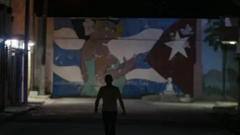 Getty Images A pedestrian walks down a street during a rolling blackout in Havana. He's in shadow but a wall mural is highlighted ahead of him. The mural features a woman in a green dress and a Cuba flag motif.