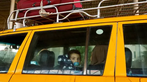 REUTERS/Ibraheem Abu Mustafa A Palestinian child, with a dummy in its mouth, looks out the window of a yellow car at the Rafah crossing in Egypt. The child is on the lap of a woman, whose face is turned away from the camera. Just visible on top of the car is a metal roof rack, with a red suitcase tied to it.