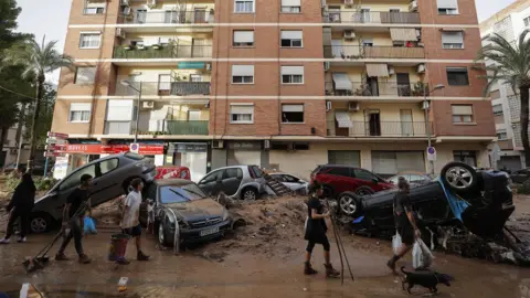 EPA People carrying buckets and brooms walk past piled-up cars along a mud-covered street