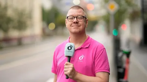 A man dressed in a pink polo shirt stands in a street, holding a microphone up in front of his chest. He wears glasses and is smiling proudly,