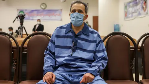 AFP Jamshid Sharmahd has short greying hair and a blue medical mask across his face, as he sits in court wearing light blue prison prison fatigues with horizontal navy stripes. A video camera and banners are visible in the background during the hearing in Tehran, in February 2022