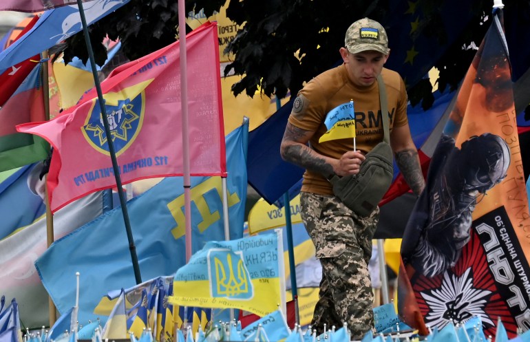 A Ukrainian soldier places a Ukrainian flag at a memorial for those killed in the war. There are lots of flags around.