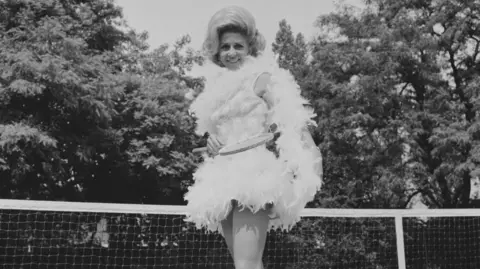 Getty Images Pericoli is seen standing in front of a tennis net, posing in a feathered outfit on court in the UK, 23rd June 1968.