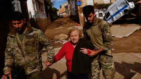 Reuters An elderly woman who looks upset is helped by two young men dressed in military fatigues. They hold her hands as she walks down a street covered in mud