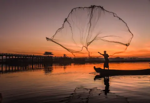 Steve Demeranville Silhouette of a man casting a large fishing net in the air from a boat during sunrise