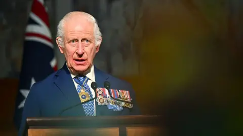 EPA King Charles delivers a speech in the Australian Parliament on Monday,  he is wearing a suit and white shirt with a blue and white tie, with several medals on his left lapel and a gold necklace around his neck. He's looking slightly away from camera, with an out-of-focus audience member in the foreground and an Australian flag in the background.