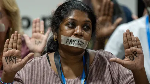 BBC A climate protestor sits with a piece of tape across her mouth with the words 'pay up!' written in black across the tape and on the palms of both hands. 