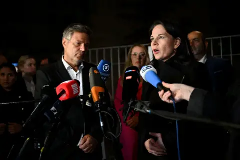 Reuters Economy Minister Robert Habeck wearing a suit and shirt and Foreign Minister Annalena Baerbock wearing a black coat stand in front of microphones to speak to the press.