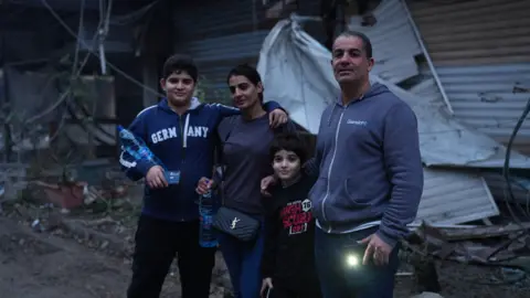 Car dealer Mohamad Marouf (R), younger son, wife and older son (L) standing in front of the destruction on their street from Israeli air strikes. Mr Marouf  is holding his mobile phone with the torch on - both sons also have phones in their hands. Older son has one arm wrapped around a water bottle, and the other around his mother's shoulders. She is also holding a water bottle.