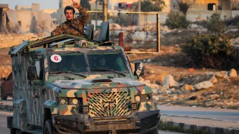 Reuters A man in a camouflage jacket stands in the back of a van driving down a road, with one hand in the air.