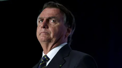 Getty Images Jair Bolsonaro, who has brown and grey combed over hair and full eyebrows, looks to the side as he stands on a lit stage while wearing a black suit with a white shirt and navy tie with yellow and green stripes