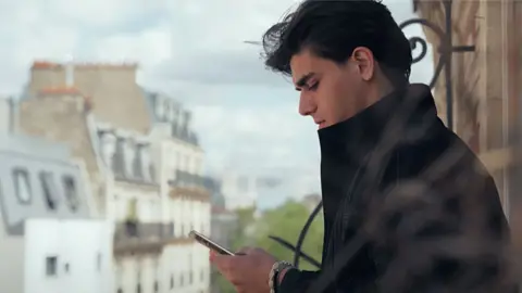 Rueben stands on a balcony in Paris with buildings in the background - he wears a long dark coat and he is photographed in profile, looking down at his phone