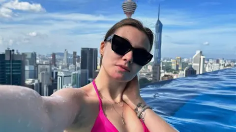 Instagram A selfie of Valeria Baigascina taken in a rooftop pool in Kuala Lumpur, with the striking skyline with tall towers behind her. Her long dark hair is tied back, she wears a pink bikini and dark sunglasses and a tattoo is visible on her wrist.