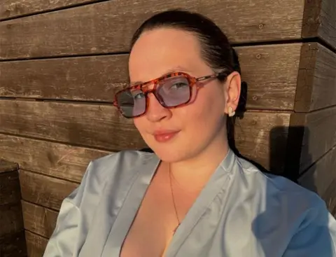 Instagram In a selfie taken at sunset, a young woman with brown hair tied back is sitting outside a wooden gite She wears a low-cut grey top, earrings and sunglasses, and is smiling at the camera. 