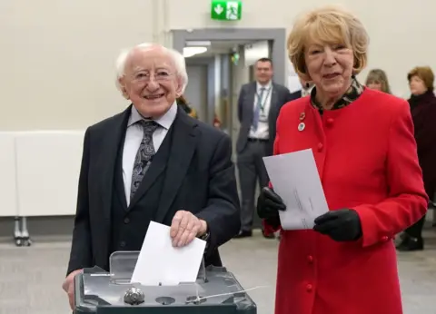 PA Michael D Higgins in black suit and white shirt, wife Sabina in red coat and black gloves, holding ballot papers and voting