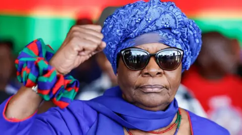 AFP Netumbo Nandi-Ndaitwah wearing dark sunglasses and a patterned blue headscarf raises her fist in a victory sign.