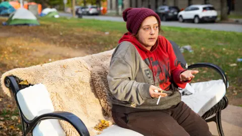 Kelly Clark/BBC Samantha Nickerson sits on a park bench in downtown Halifax. She wears a burgundy toque, a grey and red hoodie and is speaking animatedly with a cigarette in hand 